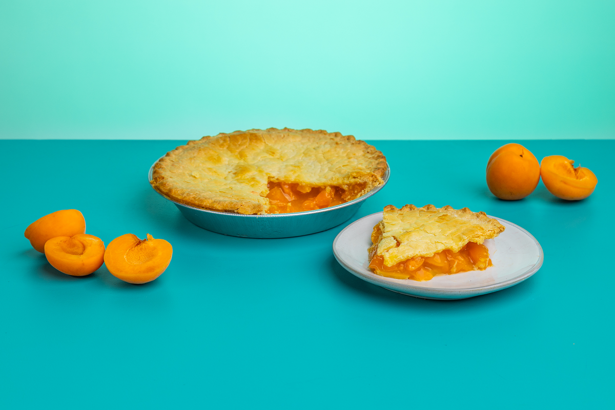 A slice of Polly's Pies Apricot pie on a plate in front of a whole Apricot pie. Apricots surround the pie in front of a blue background