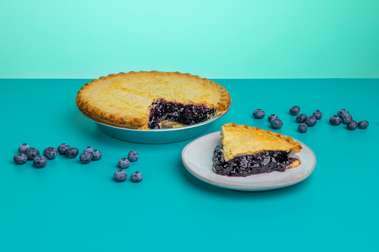 A slice of Polly's Pies blueberry pie on a plate in front of a whole blueberry pie. Blueberries surround the pie in front of a blue background