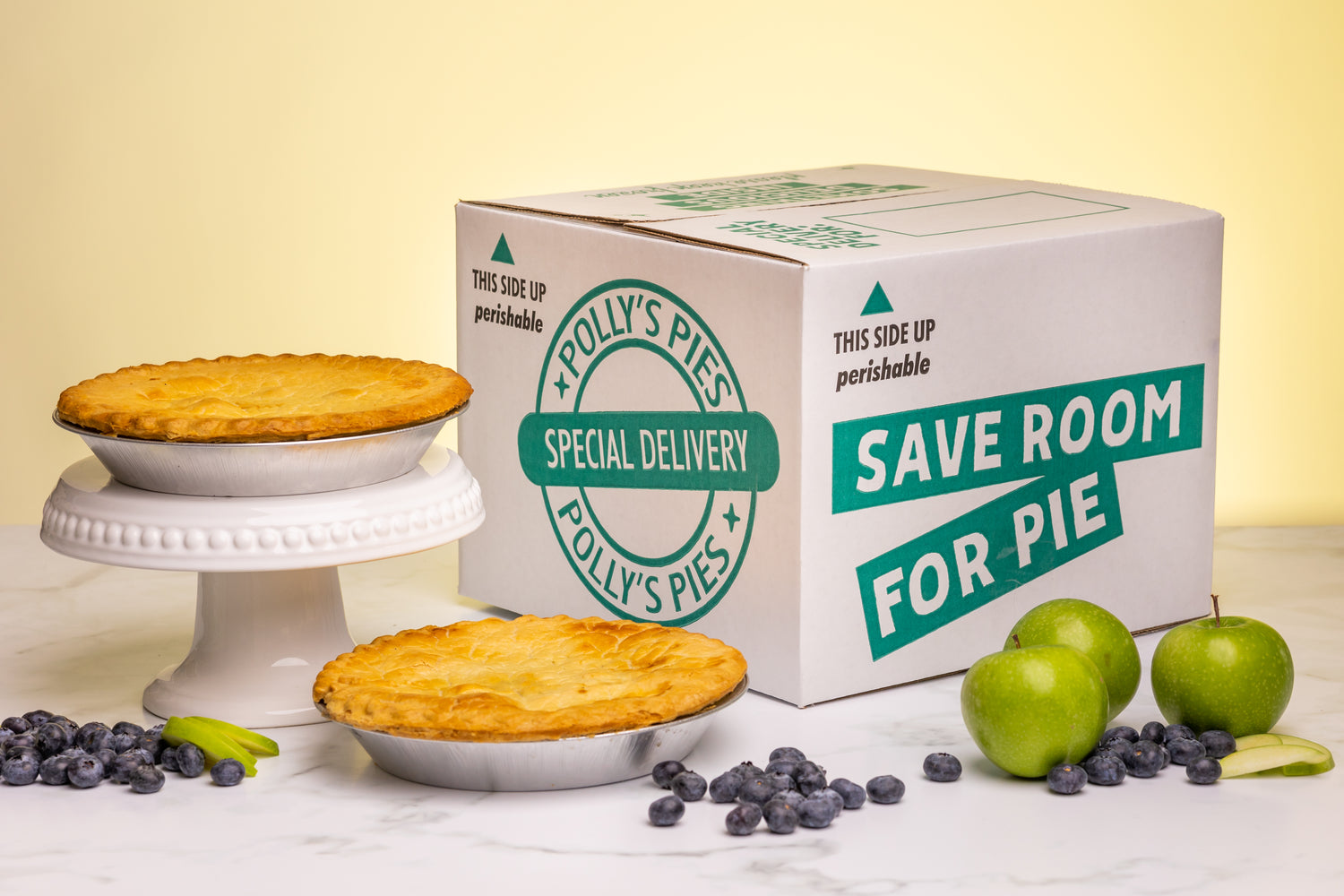 Two pies surrounded by sliced apples, whole apples, blueberries, and a Polly's Pies shipping box in front of a white marble and yellow background.