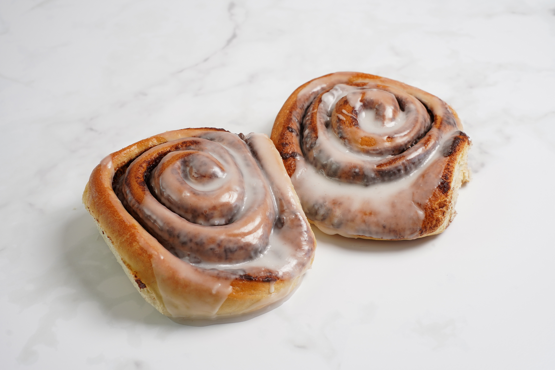 Two giant cinnamon rolls covered in icing sitting on a marble background.