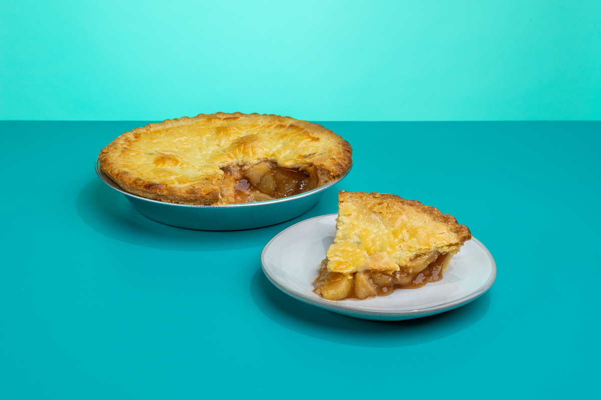 A slice of Polly's Pies apple pie on a plate in front of a whole apple pie in front of a blue background