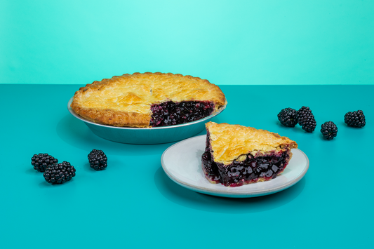 A slice of Polly's Pies boysenberry pie on a plate in front of a whole boysenberry pie. Boysenberries surrounds the pie in front of a blue background