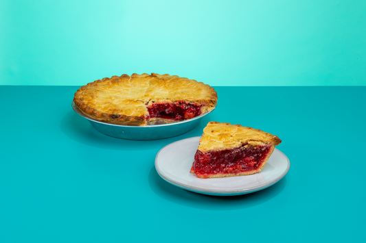 A slice of Polly's Pies Cherry pie on a plate in front of a whole cherry pie and a blue background
