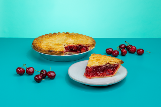 A slice of Polly's Pies cherry pie on a plate in front of a whole cherry pie. Cherries surround the pie in front of a blue background