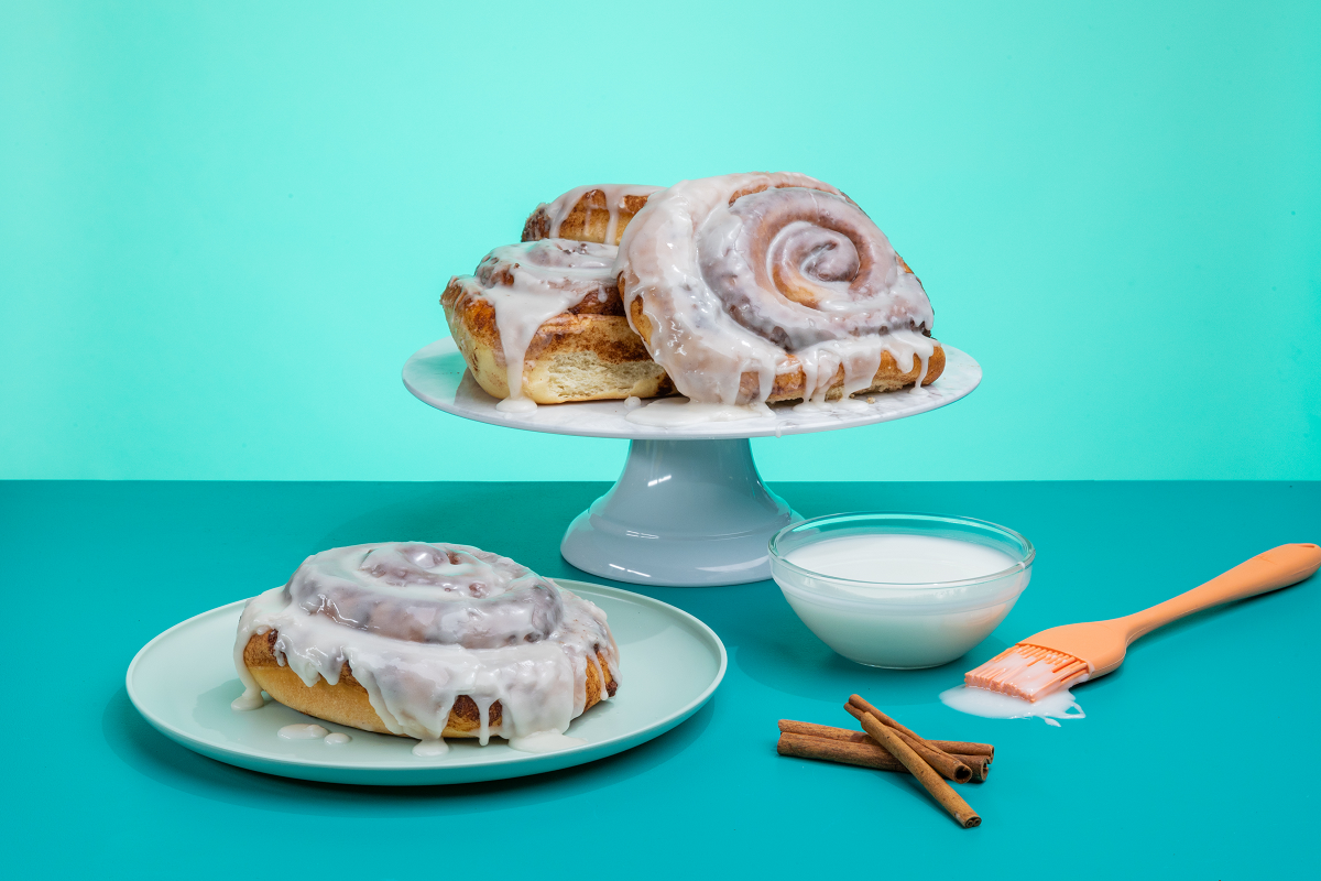 A giant cinnamon roll covered in icing on a blue plate in front of a cake stand holding three giant cinnamon rolls covered in icing. A bowl of icing, cinnamon sticks, and a pastry brush sit next to the cinnamon rolls in front of a blue background