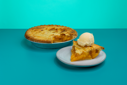 A slice of Polly's Pies peach pie on a plate topped with a scoop of ice cream in front of a whole peach pie in front of a blue background
