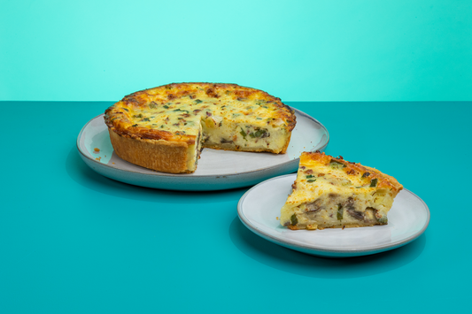 A slice of Polly's Pies quiche lorraine on a plate sitting in front of a whole quiche lorraine with a slice taken out of it in front of a blue background.