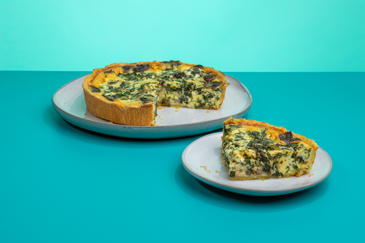A slice of Polly's Pies spinach & feta quiche on a plate sitting in front of a whole spinach & feta quiche with a slice taken out of it in front of a blue background.