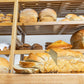 A loaf of Polly's Pies sourdough bread sliced in front of a rack of miscellaneous bakery items including dinner rolls, muffins, cornbread, and loaves of bread.