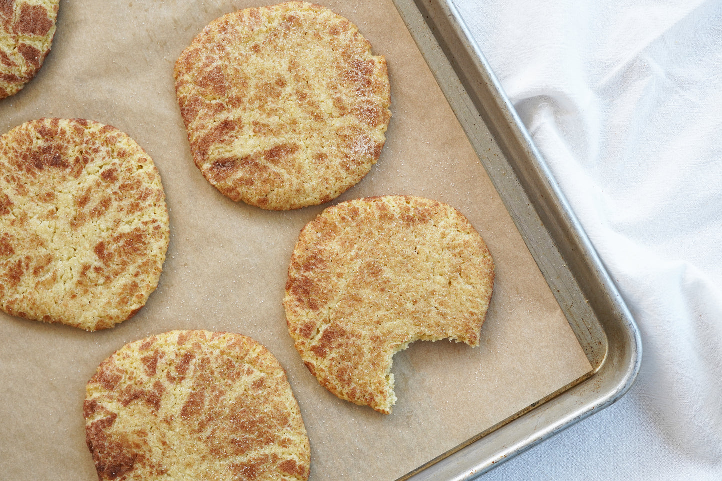 Polly's Pies snickerdoodle cookies on top of a baking sheet lined with parchment paper. One cookie has a bite taken out if it.