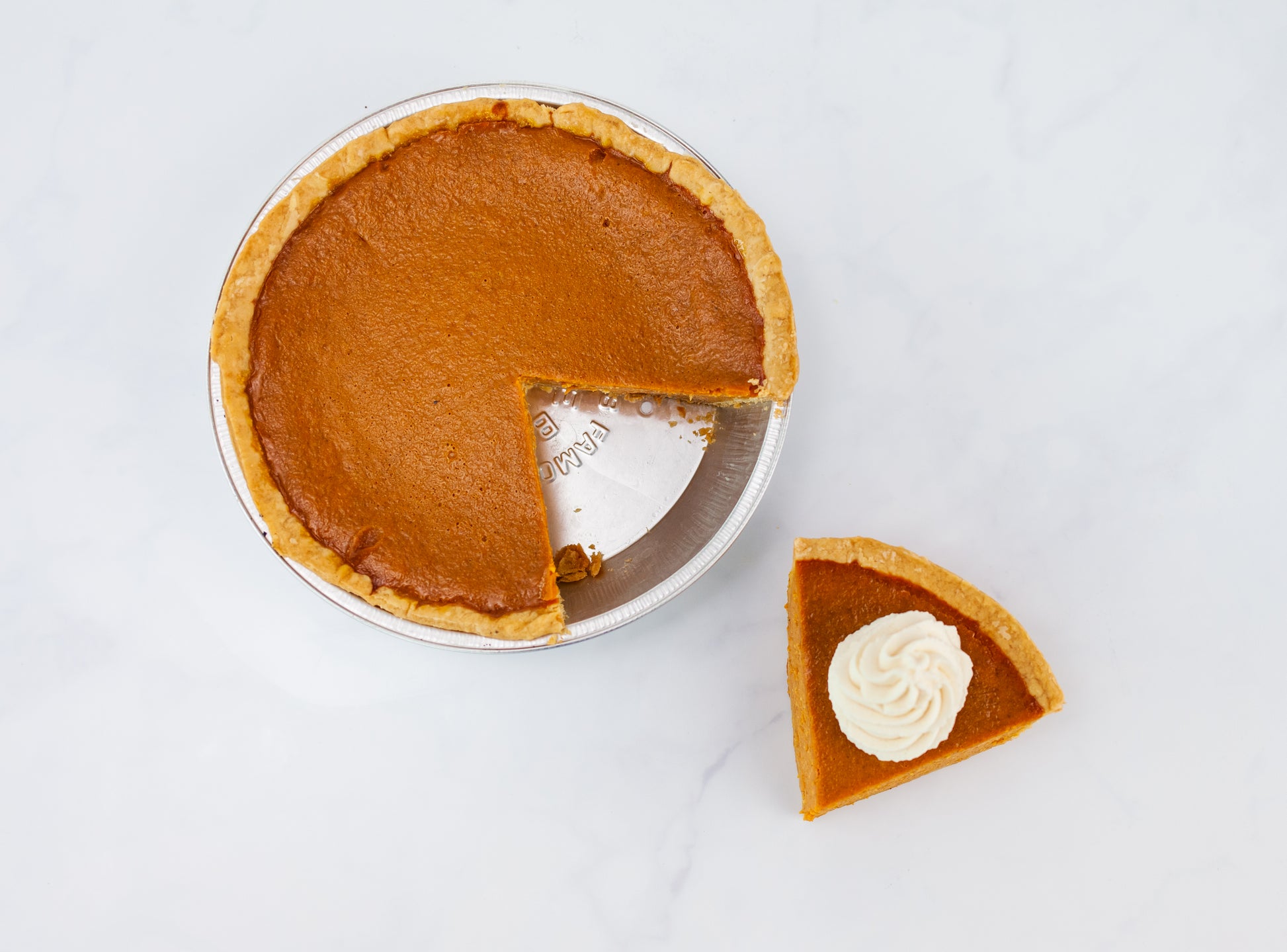 A whole Polly's pumpkin pie with a slice taken out of it sitting by a slice of Pumpkin pie topped with a dollop of real whipped cream on a white marble background.