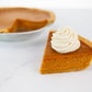 A slice of Polly's Pies pumpkin pie topped with a dollop of whipped cream with a whole pumpkin pie in the background.