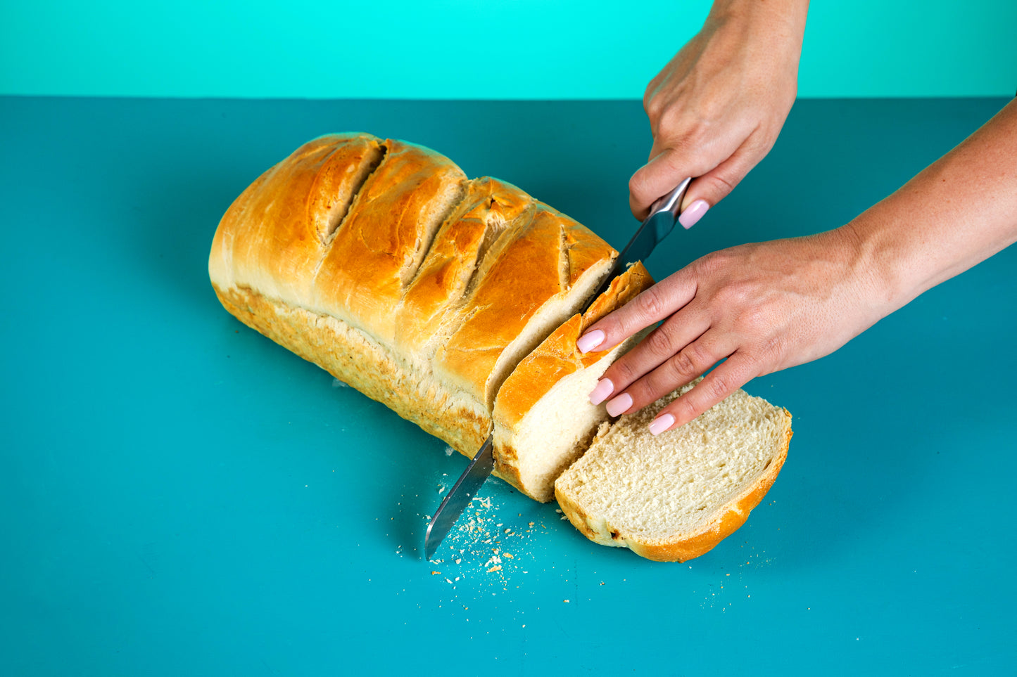 Polly's Pies loaf of Sourdough Bread on top of  a blue background. A hand  with pink nail polish is slicing the loaf with a bread knife.