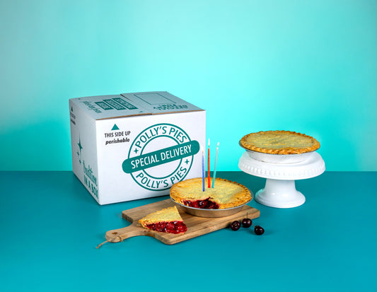 A Polly's Pies shipping box in front of a blue background. Sitting in front is a cherry pie with birthday candles and a slice taken out. There is a Chicken pie on a pie stand next to that.