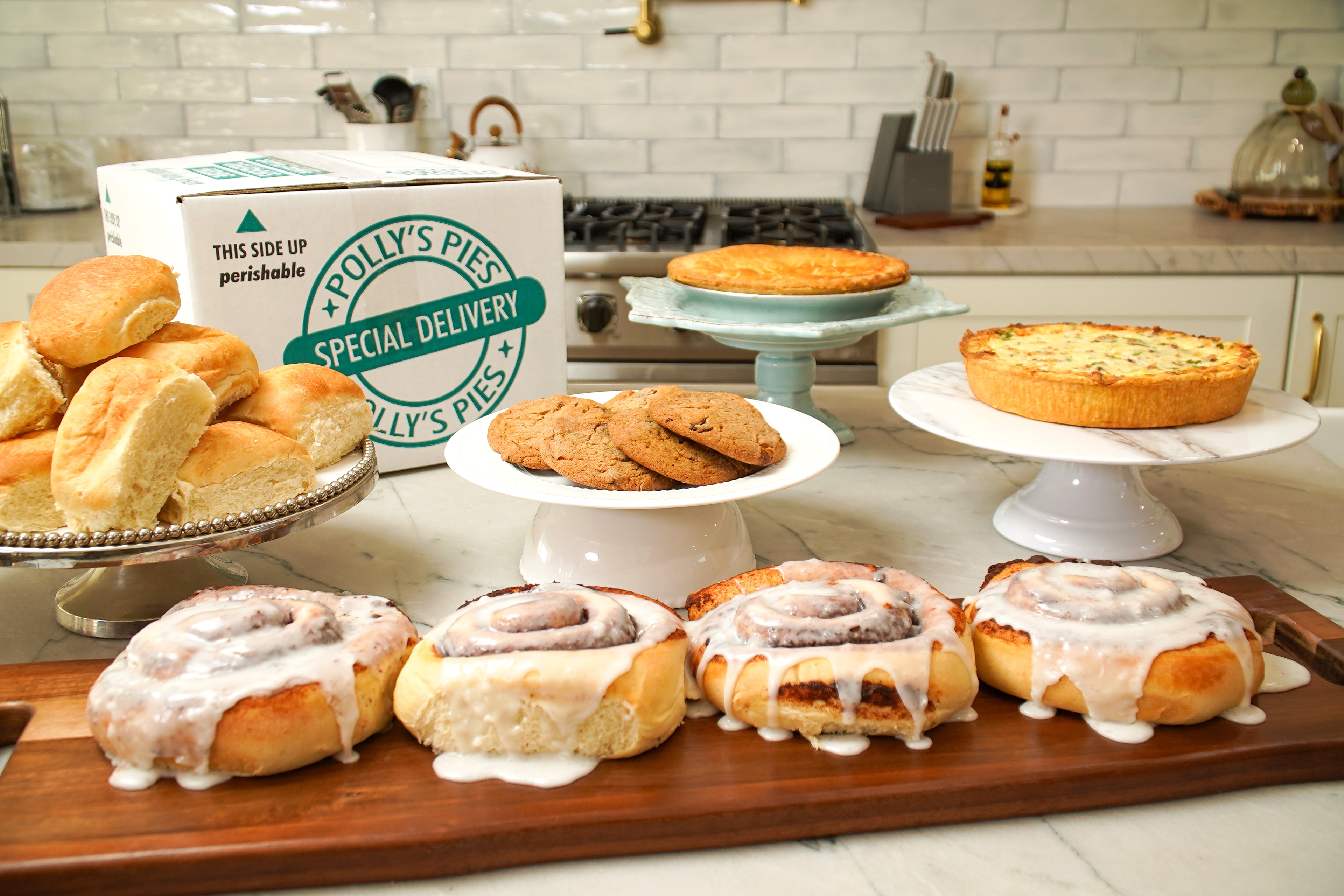 Polly's Pies cinnamon rolls, dinner rolls, cookies, quiche, pie, and delivery box sitting on a marble kitchen countertop in a white kitchen.